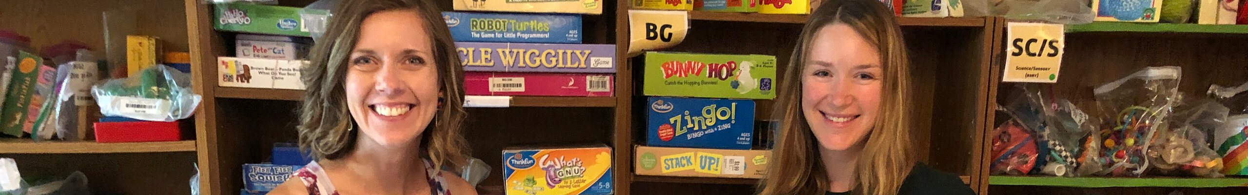 Two volunteers smiling in front of the toy library's game shelves